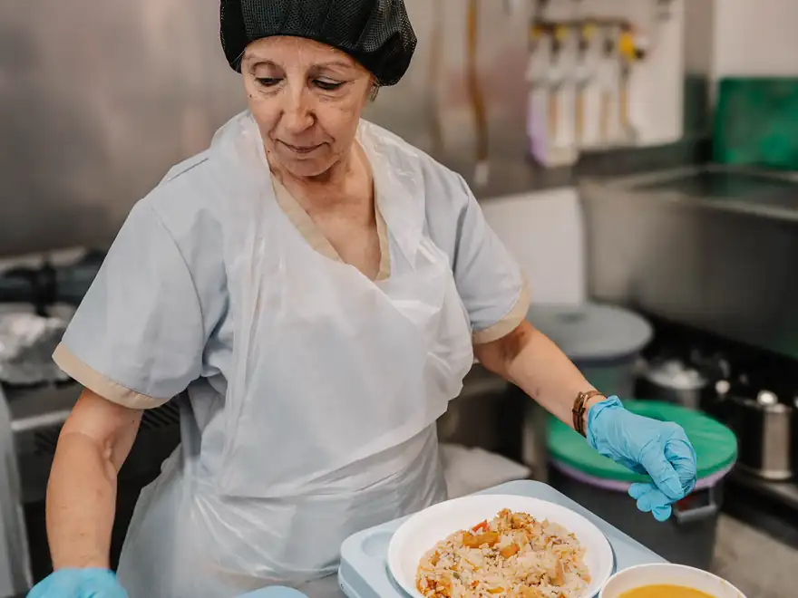 Uma cozinheira monta o tabuleiro da refeição, com prato, sopa e fruta.