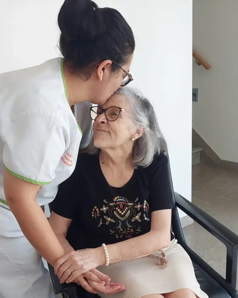 Uma auxiliar da associação, inclinada, dá um beijo na testa a uma senhora idosa sentada numa cadeira, enquanto lhe segura a mão.
